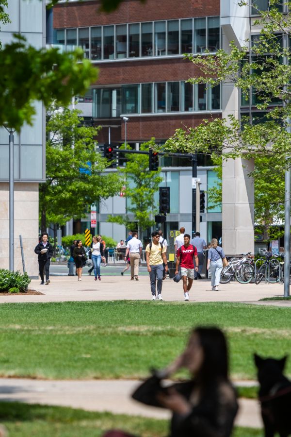 A busy campus walkway with students walking in various directions between modern buildings and green spaces, with bicycles parked nearby