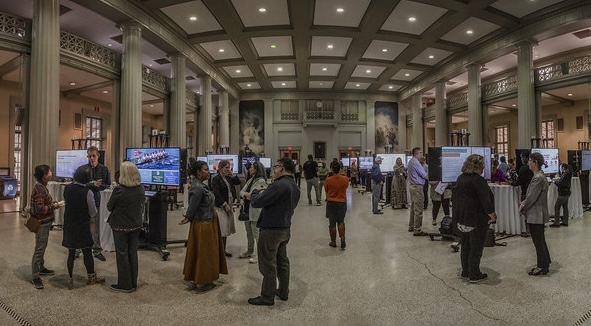 People networking and viewing digital displays in a large hall with columns and high ceilings.
