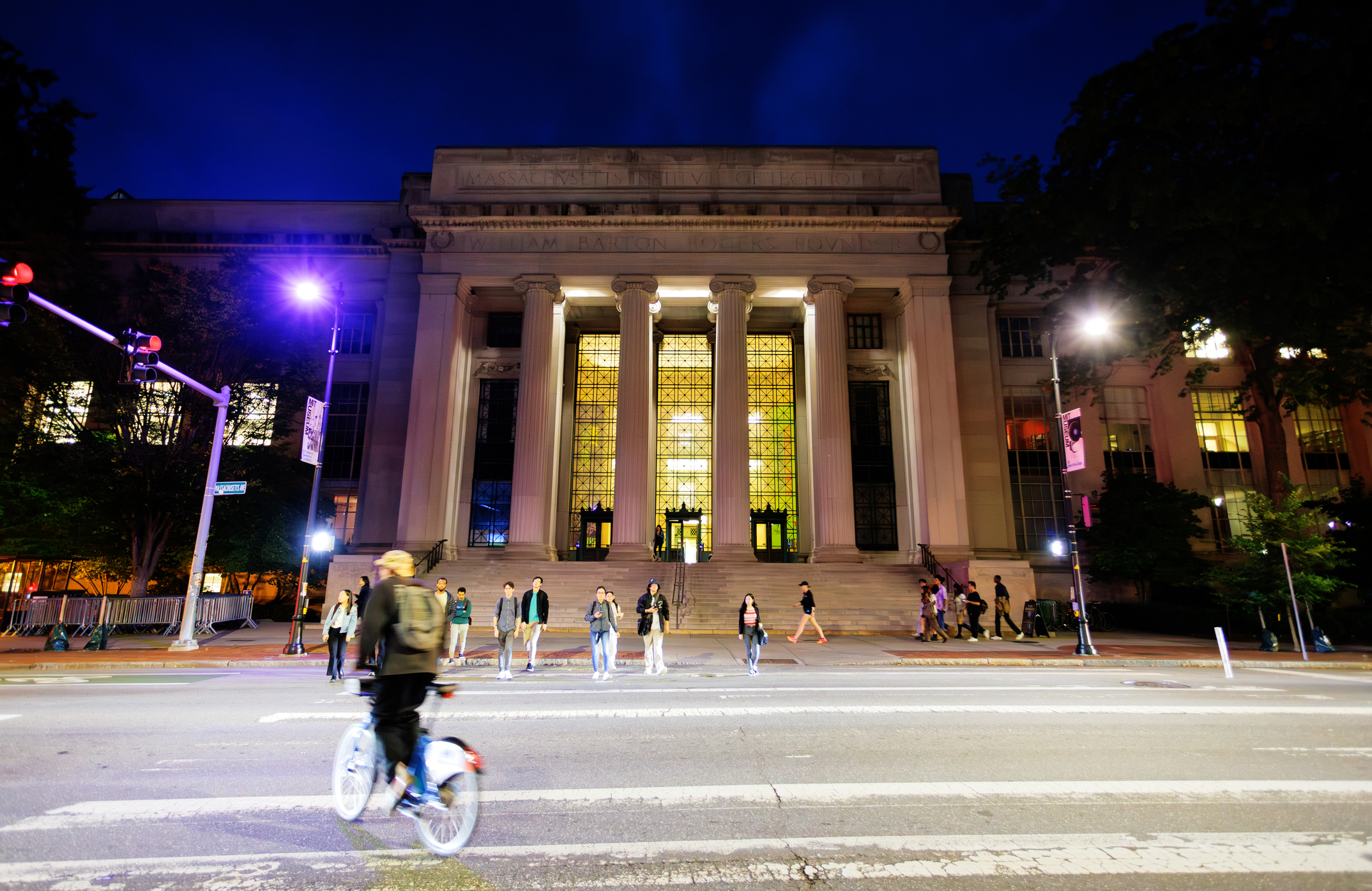People crossing a street in front of a large columned building lit up at night.